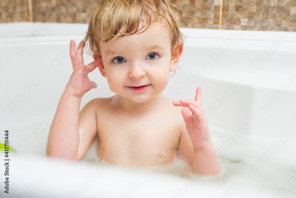 Funny little boy getting a bath in the bathroom. A child bathes in water with soap suds in white bath Funny little boy getting a bath in the bathroom. A child bathes in water with soap suds in white bath