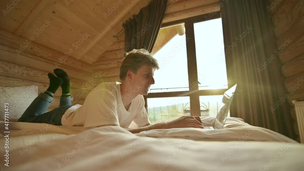 Positive young man lying in the apartment bed and working online on a laptop with a smile on his face. Work online in a hotel room on a laptop. Positive young man lying in the apartment bed and working online on a laptop with a smile on his face. Work online in a hotel room on a laptop.