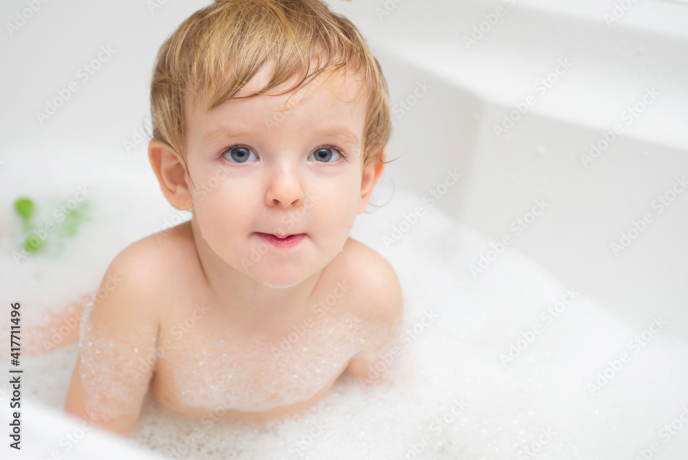 Funny little boy getting a bath in the bathroom. A child bathes in water with soap suds in white bath Funny little boy getting a bath in the bathroom. A child bathes in water with soap suds in white bath