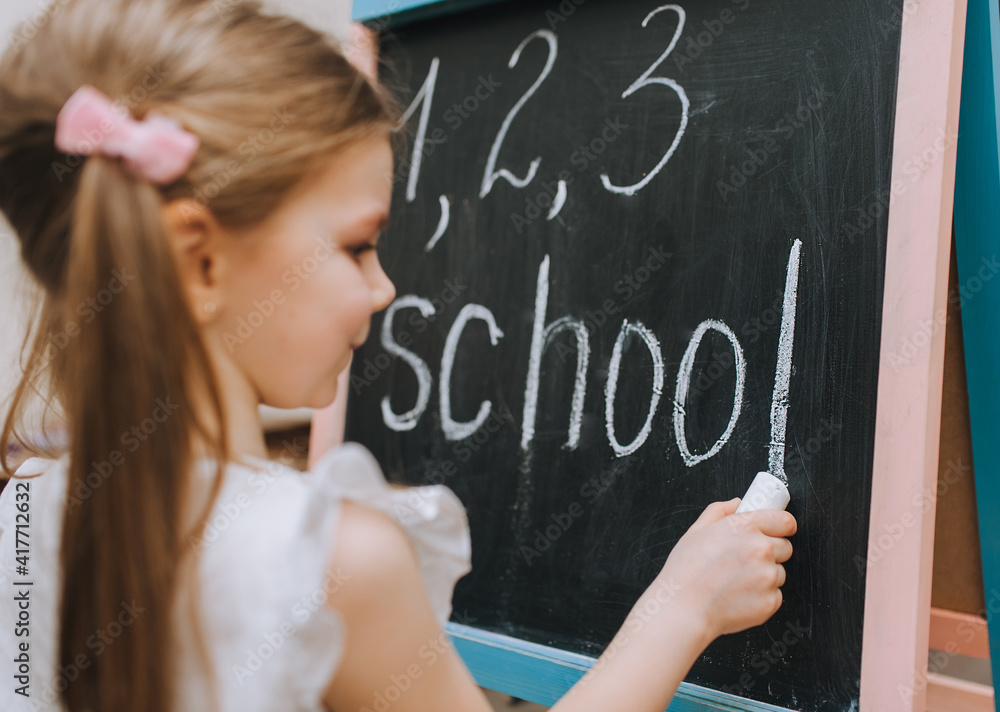 A red-haired girl, a child with pigtails, writes diligently, draws on a black wooden board, an easel, holding white chalk, the word school and numbers in her hand. Photography, copy space, concept. A red-haired girl, a child with pigtails, writes diligently, draws on a black wooden board, an easel, holding white chalk, the word school and numbers in her hand. Photography, copy space, concept.