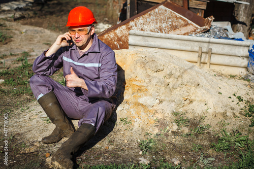 A young construction worker in an orange helmet and protective glasses sits on a pile of sand. Building. Portrait. The builder is talking on the phone.
