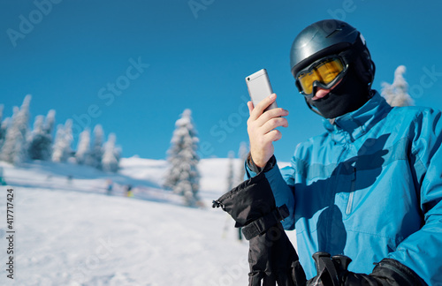 Happy man using smartphone in snowy mountains after skiing. Guy texting phone message. Cheerful sporty skier enjoy winter vacation at mountain with cellphone.