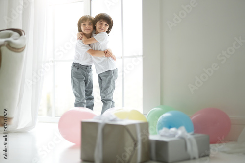 Best friends. Two cute latin twin boys, little children in casual wear hugging each other, looking happy, standing in the room with colorful balloons and giftboxes