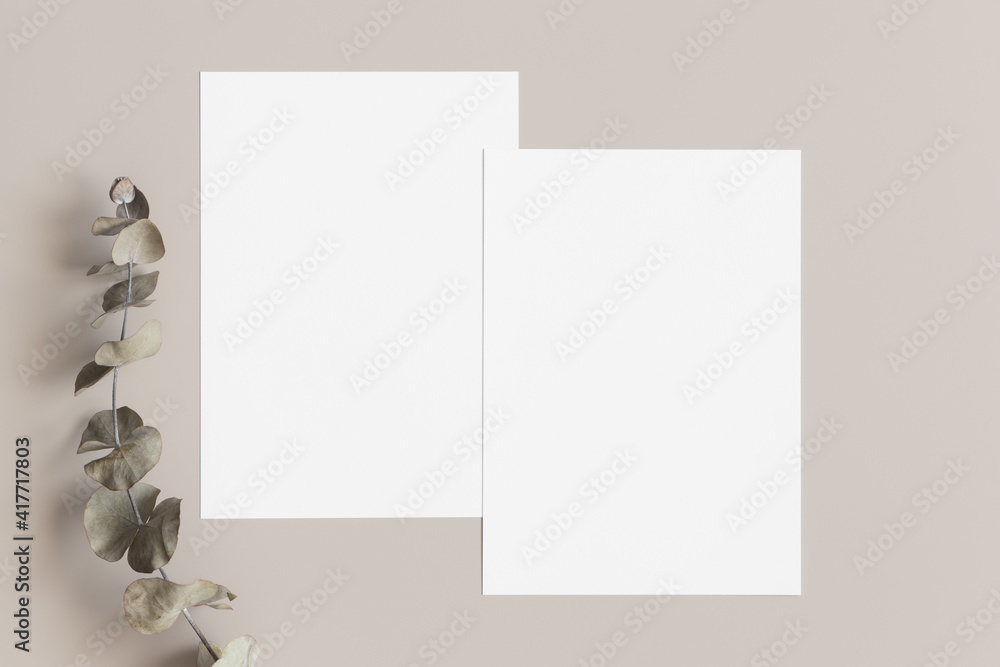 Two white invitation cards mockup with an eucalyptus decoration on the beige table. 5x7 ratio, similar to A6, A5. Two white invitation cards mockup with an eucalyptus decoration on the beige table. 5x7 ratio, similar to A6, A5.