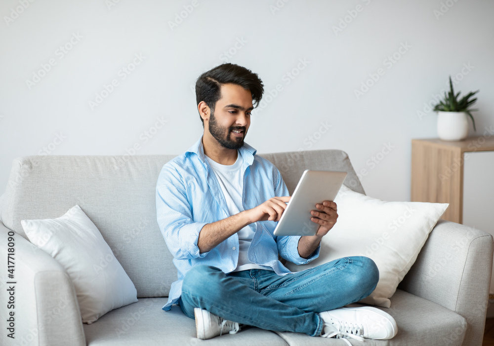 Eastern Millennial Guy Spending Time At Home With Digital Tablet Eastern Millennial Guy Spending Time At Home With Digital Tablet
