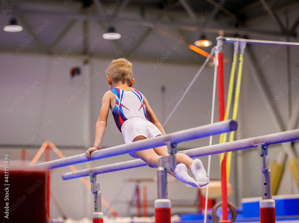 Naklejka premium A little boy in sportswear performs an exercise on the uneven bars. Rhythmic gymnastics, training in the hall.