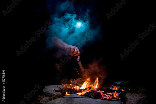 night view of bonfire with saucepan and man's hand stirring dish in it
