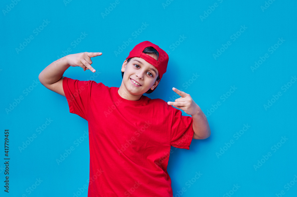 Portrait of boy in t-shirt and red cap putting the horns with his fingers, on blue background. Rapper gesture. Copy space Portrait of boy in t-shirt and red cap putting the horns with his fingers, on blue background. Rapper gesture. Copy space