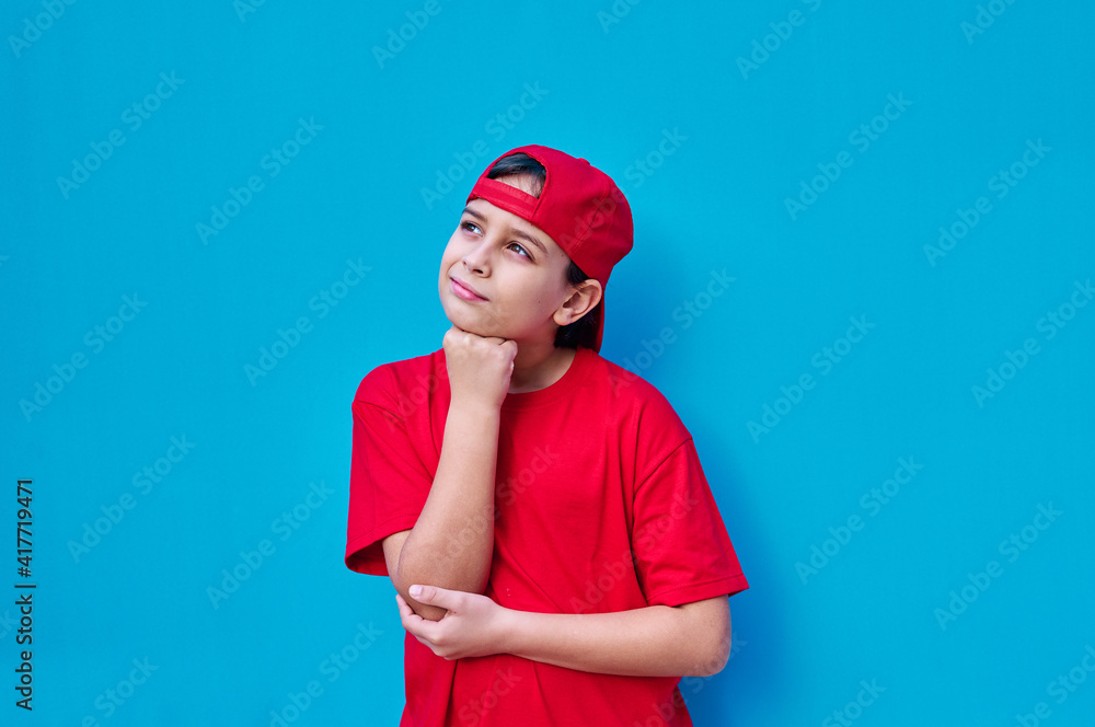 Portrait of pensive boy in red cap and t-shirt with hand on face, on blue background. Copy space Portrait of pensive boy in red cap and t-shirt with hand on face, on blue background. Copy space