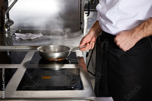 Cropped chef at kitchen restaurant. Cook in white uniform professionally preparing meal on stove, fries a dish for guests. close-up photo, side view