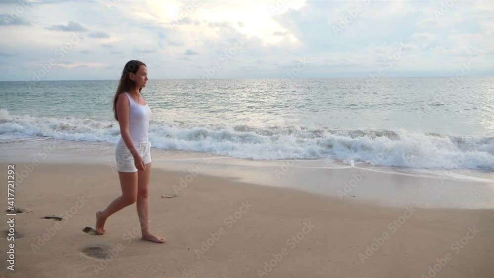 Relaxed woman stroll along sea beach at evening hour, slow motion trucking shot. She wear white summer clothes, walk alone and enjoy sound of breaking waves Relaxed woman stroll along sea beach at evening hour, slow motion trucking shot. She wear white summer clothes, walk alone and enjoy sound of breaking waves