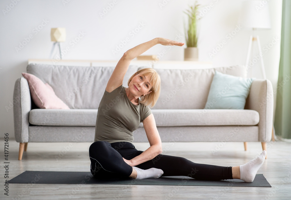 Full length portrait of athletic mature woman stretching her leg on yoga mat in living room, copy space Full length portrait of athletic mature woman stretching her leg on yoga mat in living room, copy space