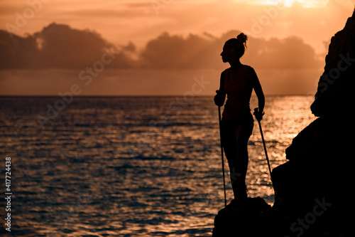 awesome view of female silhouette with trekking sticks near cliff on background with sunrise