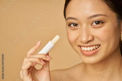 Face closeup of adorable young woman with beautiful smile looking at camera and holding lip balm, posing isolated over beige background
