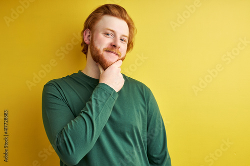 european male with red hair and beard in green shirt posing at camera isolated in studio, male stands touching chin. people concept