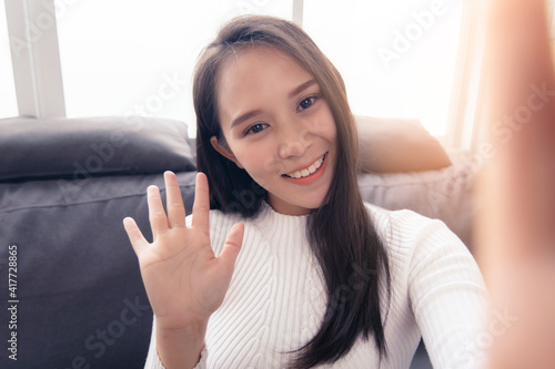 Happy smiling millennial young Asian woman greeting waving hands self portrait holding camera view, at home video blog vlogging self recording, social media influencer content creator lifestyle