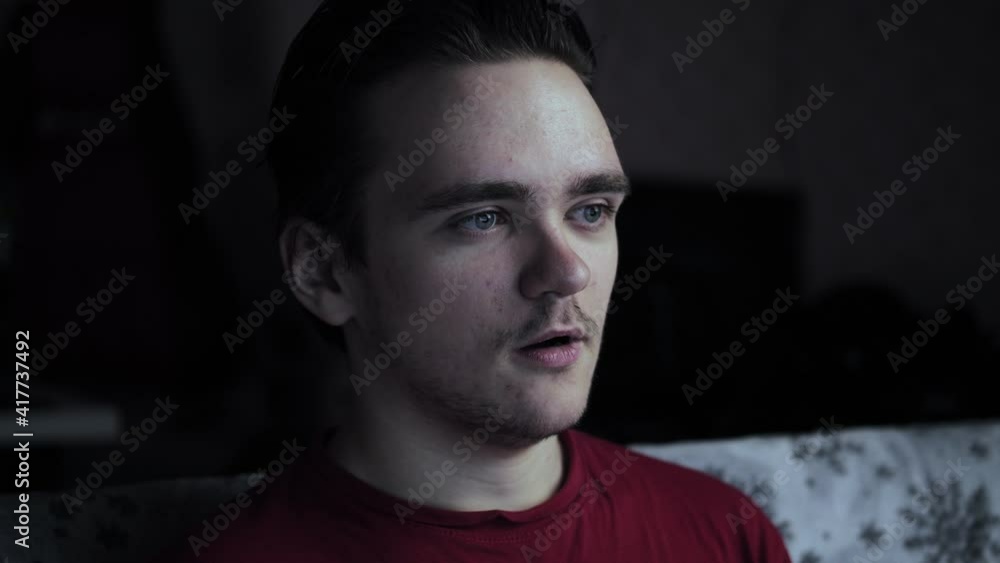 A young guy sits on the couch in a dark room, watches TV and is surprised at what is there. Weekend and relaxation concept