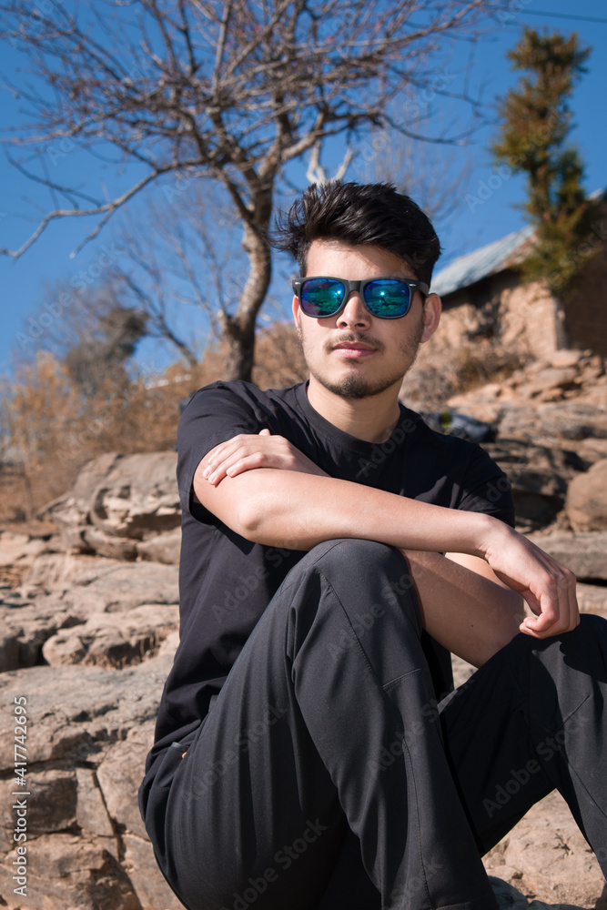 boy model sitting on rocks wearing black t-shirt black pant, sunglasses and looking at camera boy model sitting on rocks wearing black t-shirt black pant, sunglasses and looking at camera