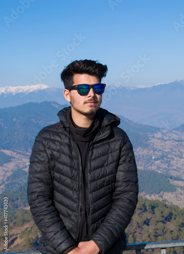 Indian ethnicity boy wearing black jacket and blue sunglasses looking at camera standing on top of mountain