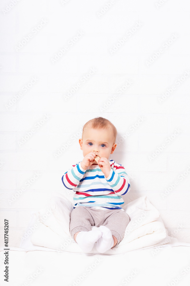Funny caucasian baby boy in striped t-shirt sitting on pillow mountain with cookie in hand. White brick wall background with copy space. Funny caucasian baby boy in striped t-shirt sitting on pillow mountain with cookie in hand. White brick wall background with copy space.