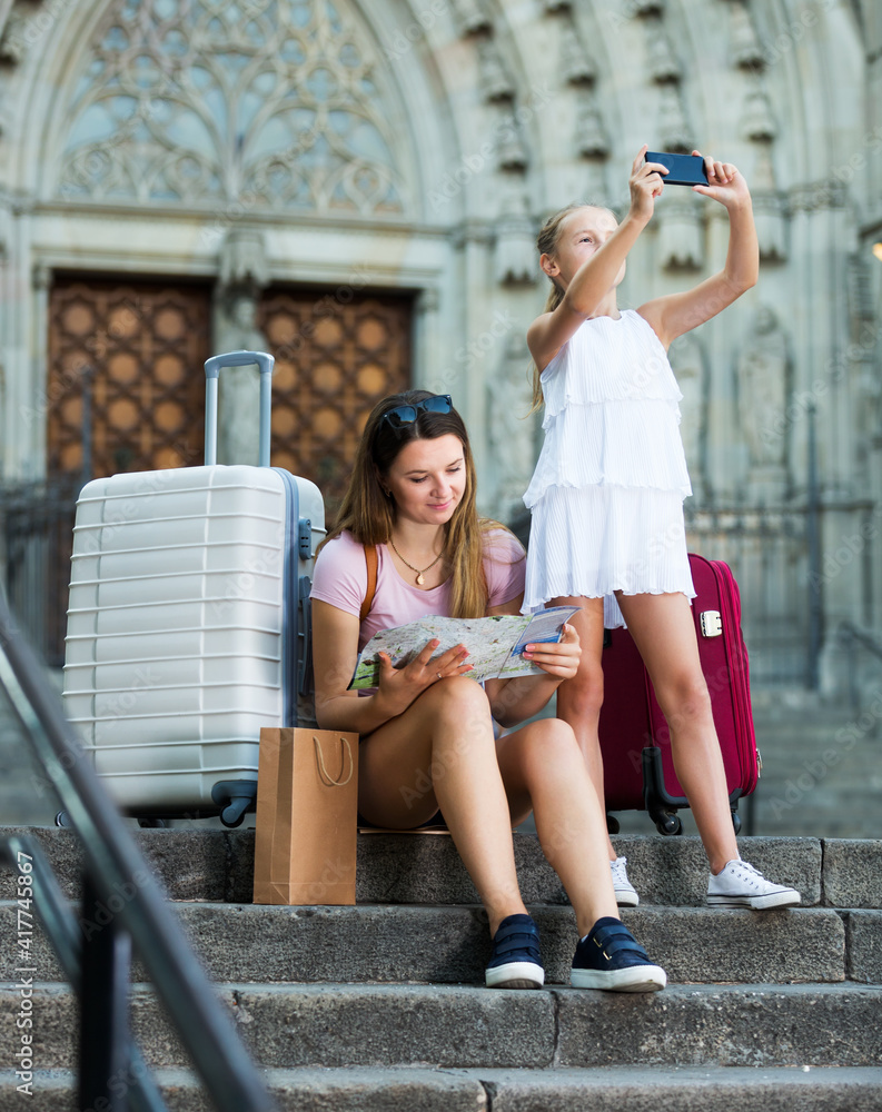 Girl taking pictures of city with phone, woman sitting on stone steps with map