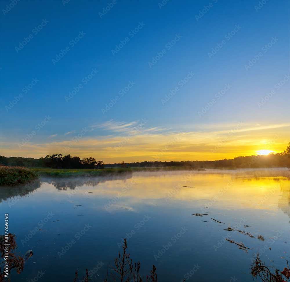 Fototapeta premium Serene lake at sunrise in forested, sun reflecting in the water with fog