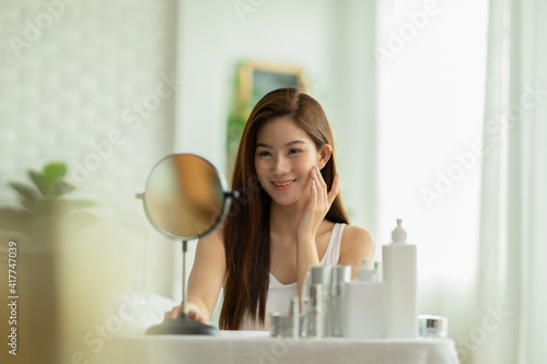 Happy Asian young woman smile and looking in mirror doing skin care touch cheek and beauty treatment at home,Wellness woman self care with beauty product at home