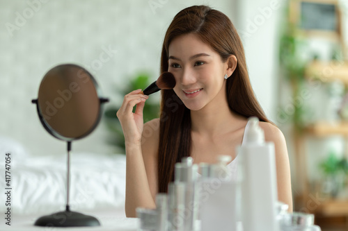 Happy Asian young woman smile and looking in mirror doing skin care applying make up brush on her cheek and beauty treatment at home Wellness woman self care with beauty product at home