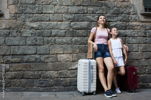 Smiling adult mother and daughter with travelling bag on stone wall background
