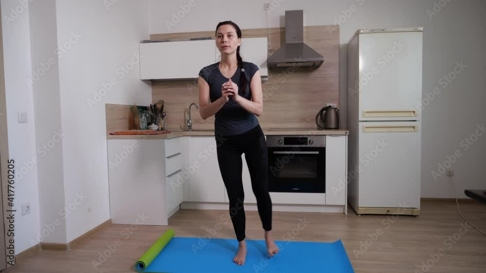 A young brunette girl training at home performs an exercise of waving her feet while standing on a blue gymnastics mat at home in the kitchen. Home workout video. High quality FullHD footage