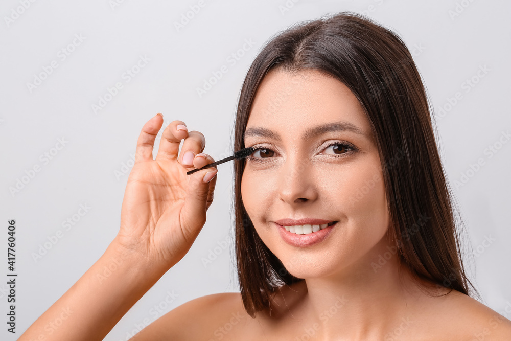 Beautiful young woman applying mascara against light background Beautiful young woman applying mascara against light background