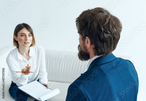A man in a blue jacket and a beard talks to a woman on the sofa indoors