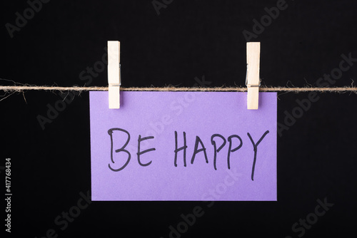  Be Happy word written on a Purple color sticky note hanging with a wire on black background