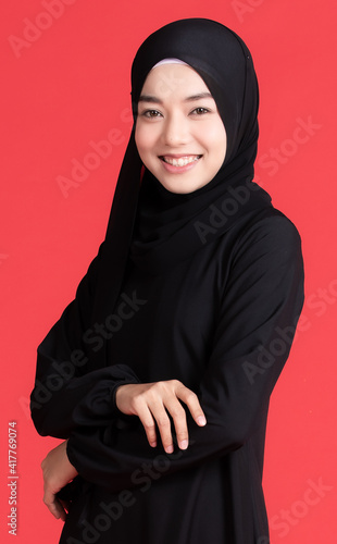 Beautiful Muslim Asian woman wearing black hijab, standing pose and smiling cheerful on red background. In religious rituals  concept
