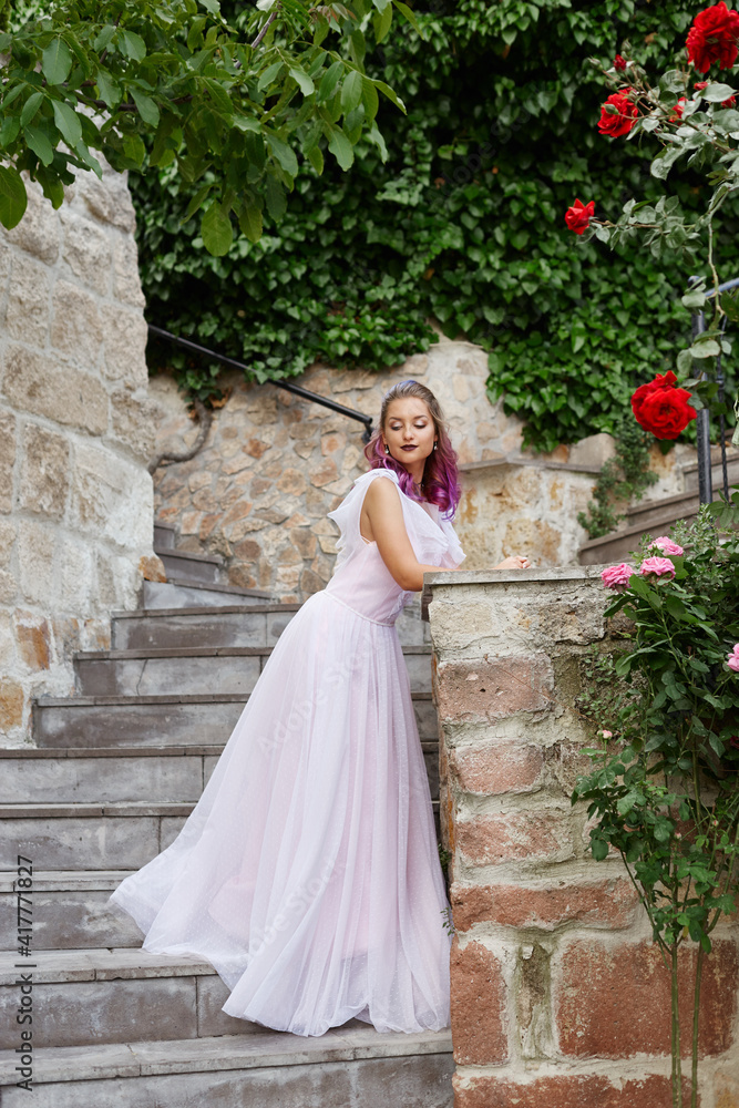 Fototapeta premium Beautiful woman with purple hair in a white wedding dress walks in the garden. Natural makeup