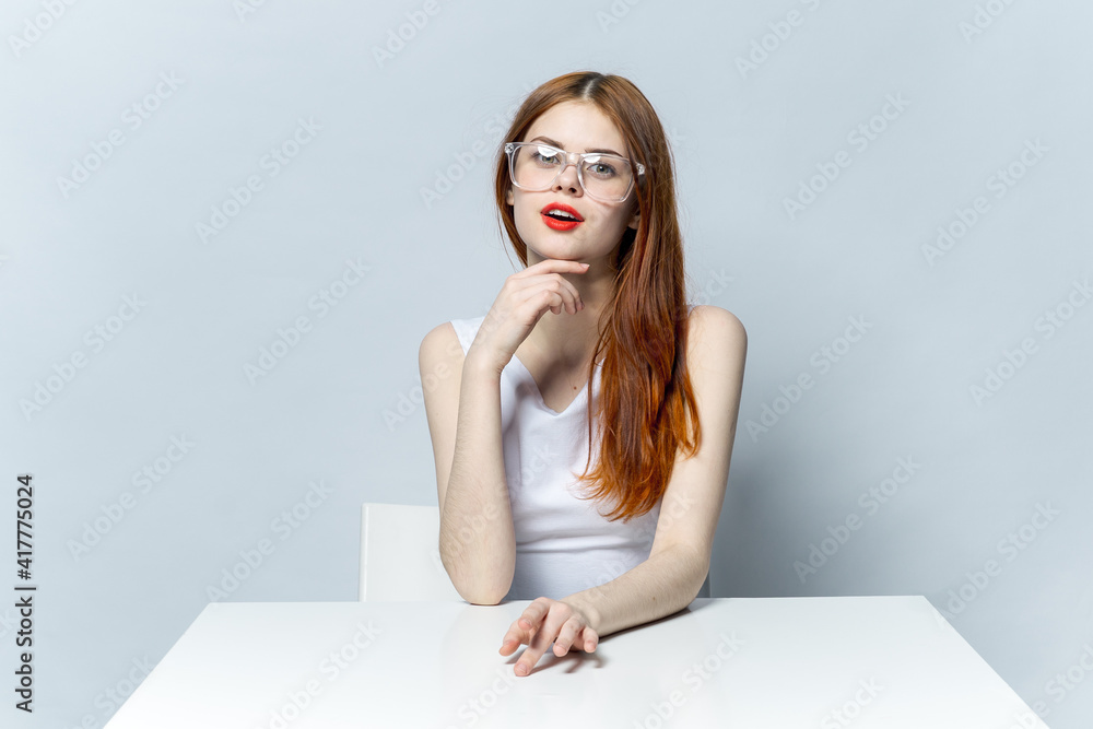 pretty red-haired woman sitting at the table wearing glasses red lips smile light background pretty red-haired woman sitting at the table wearing glasses red lips smile light background