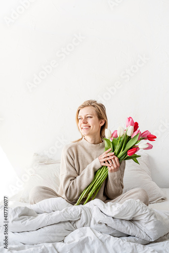 Happy woman sitting on the bed wearing pajamas holding tulip flowers bouquet