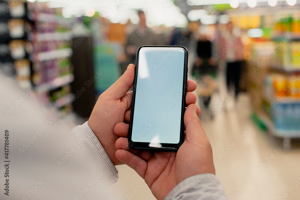 Mock-up of a smartphone with a white screen in the hands of a man. Phone on the background of in a supermarket. Mock-up of a smartphone with a white screen in the hands of a man. Phone on the background of in a supermarket.