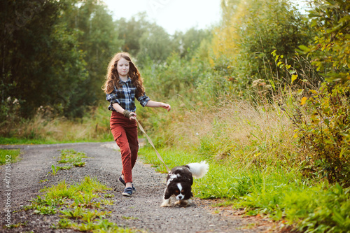 happy kid girl walking with her cavalier king charles spaniel dog on summer country road. Training her puppy and having fun.