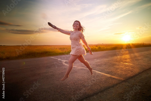 Beautiful sexy girl in pink dress and with high-heeled shoes in hand walking on the asphalt highway during sunset and field with grass on the side of the road