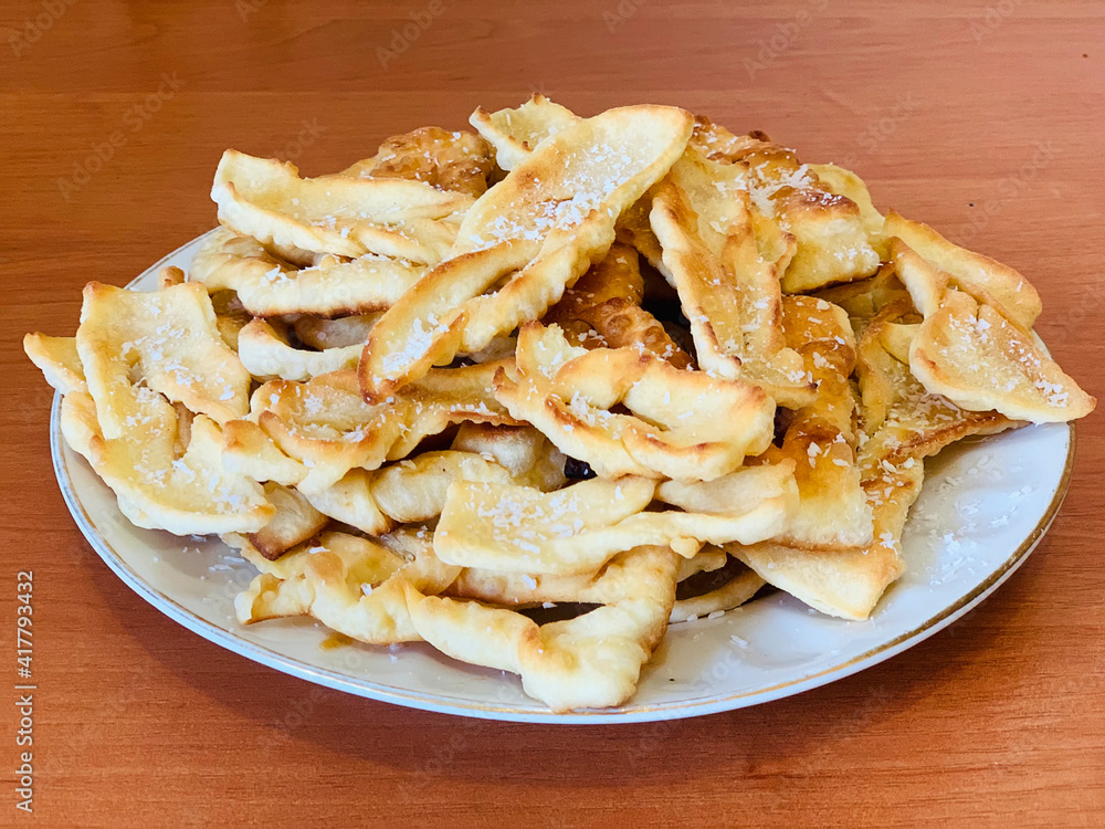 Brushwood - crispy deep fried cookies in powdered sugar on the white plate and wooden table. A traditional treat at European festivals. Homemade cookies, dessert Brushwood - crispy deep fried cookies in powdered sugar on the white plate and wooden table. A traditional treat at European festivals. Homemade cookies, dessert