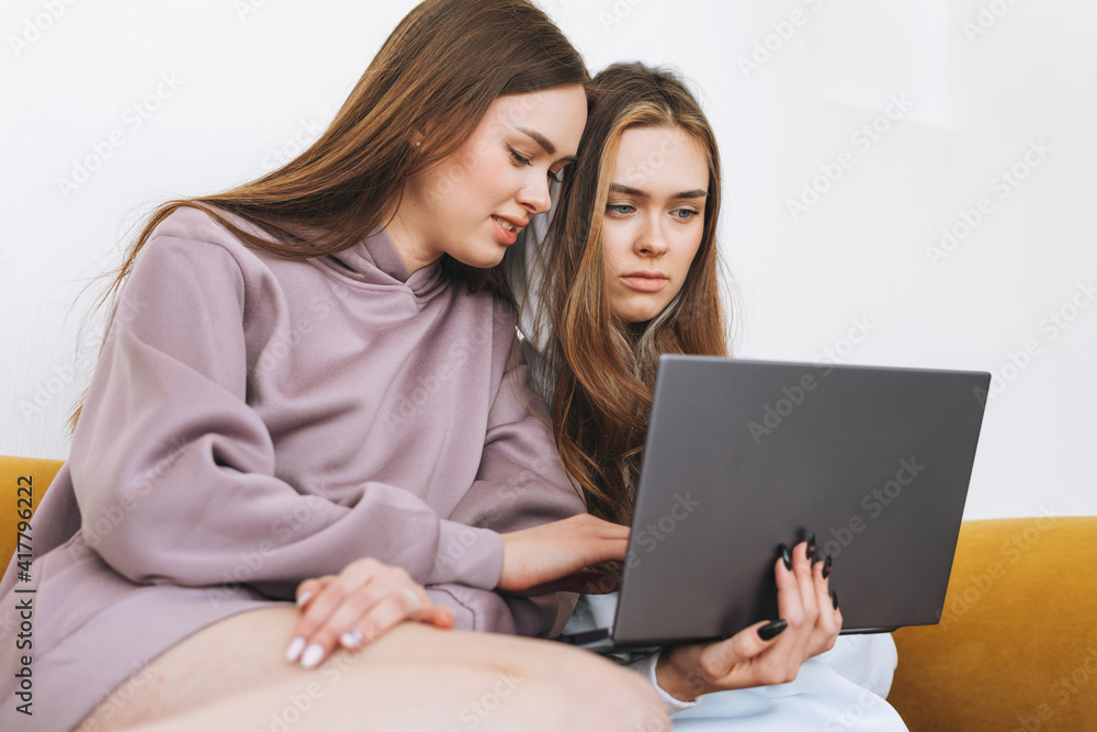 Young women sisters twins teenagers in hoodie beautiful girls using laptop sitting on yellow couch in modern interior