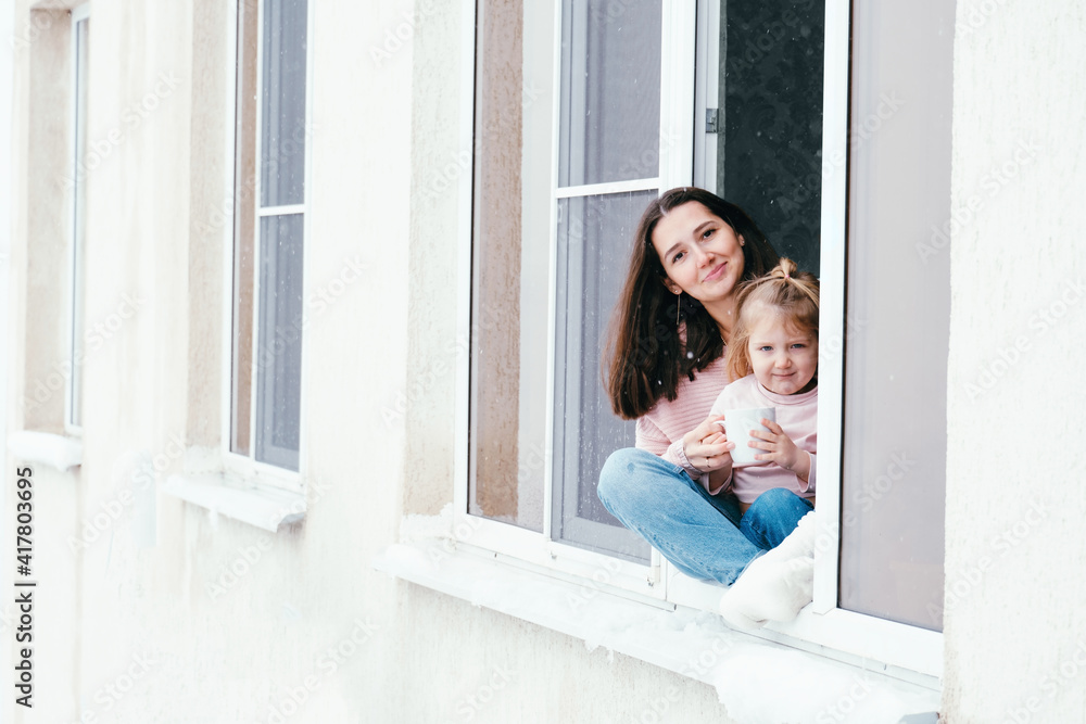 Cute portrait of mother and daughter sitting on a window sill at home with cup of hot drink. Cute portrait of mother and daughter sitting on a window sill at home with cup of hot drink.