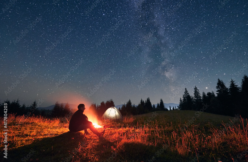 Silhouette of male traveler sitting near campfire under night sky with stars and Milky way. Starry sky under grassy hill with hiker, bonfire and camp tents. Concept of travelling and night camping. - obrazy, fototapety, plakaty 