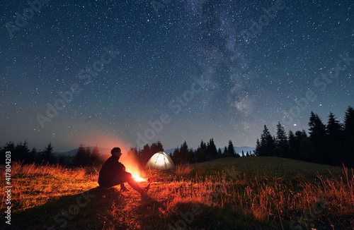 Silhouette of male traveler sitting near campfire under night sky with stars and Milky way. Starry sky under grassy hill with hiker, bonfire and camp tents. Concept of travelling and night camping.