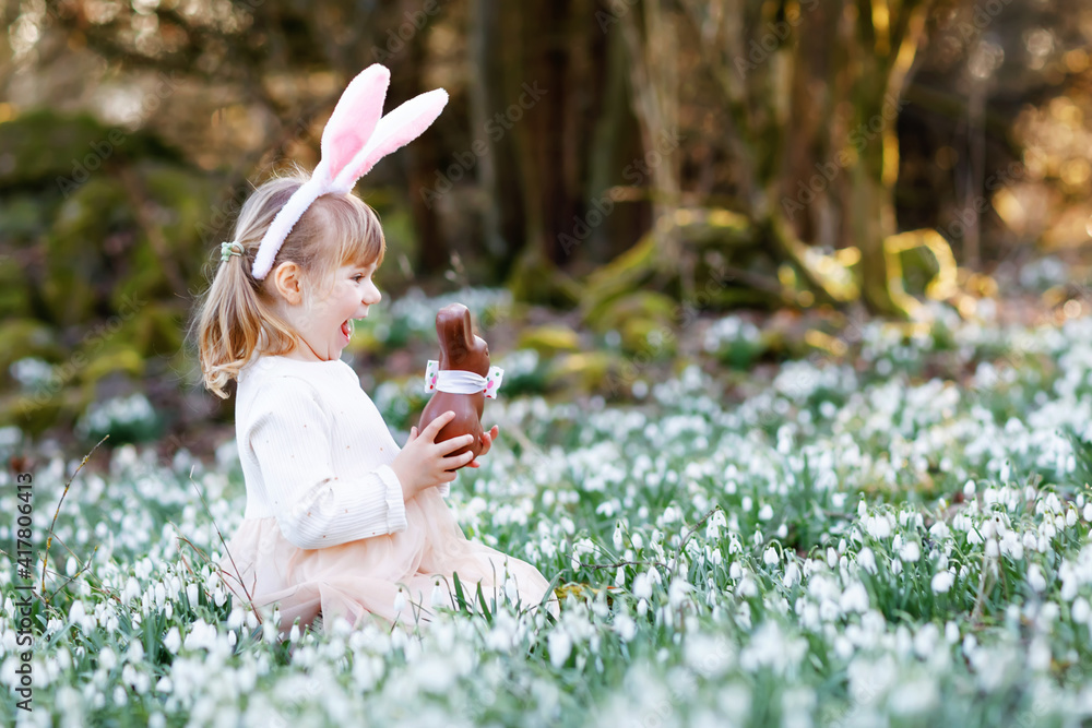 Adorable little girl with Easter bunny ears eating chocholate figure in spring forest on sunny day, outdoors. Cute happy child with lots of snowdrop flowers. Springtime, christian holiday concept. Adorable little girl with Easter bunny ears eating chocholate figure in spring forest on sunny day, outdoors. Cute happy child with lots of snowdrop flowers. Springtime, christian holiday concept.