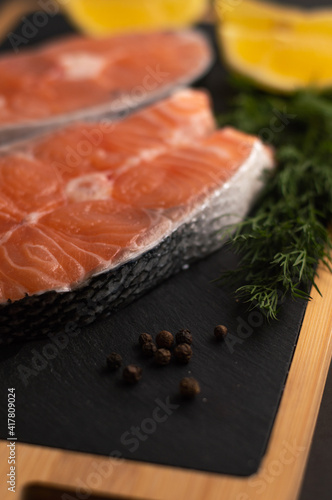 Blurred image of salmon steak, lemon wedges, herbs and peppercorns on a dark board.