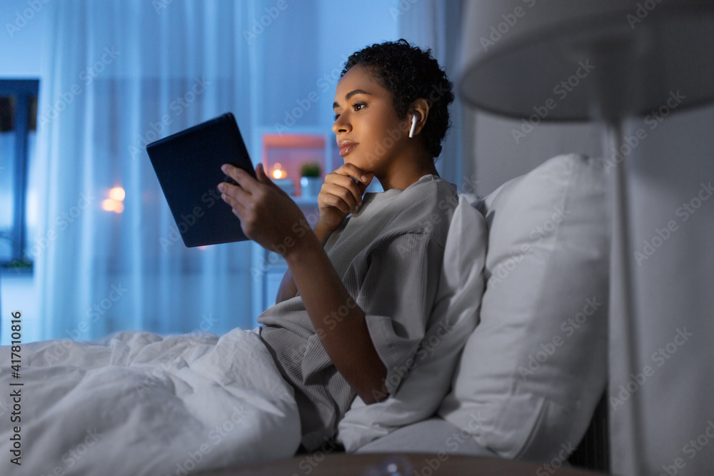 technology, internet and people concept - young african american woman with tablet pc computer and wireless earphones lying in bed at home at night technology, internet and people concept - young african american woman with tablet pc computer and wireless earphones lying in bed at home at night