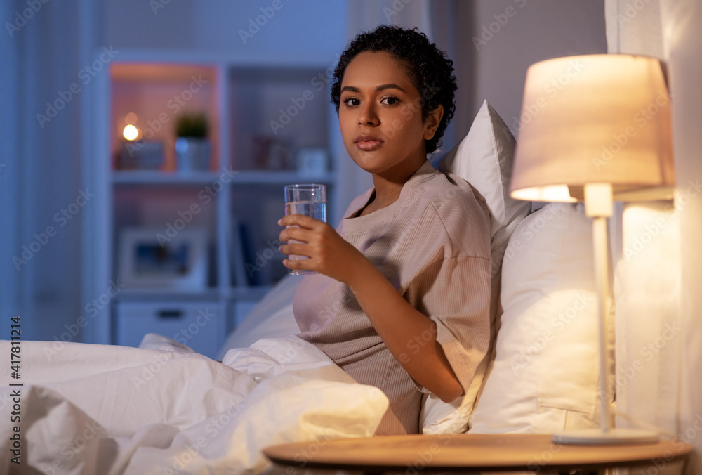 people, bedtime and thirst concept african american woman drinking water in bed at night Stock