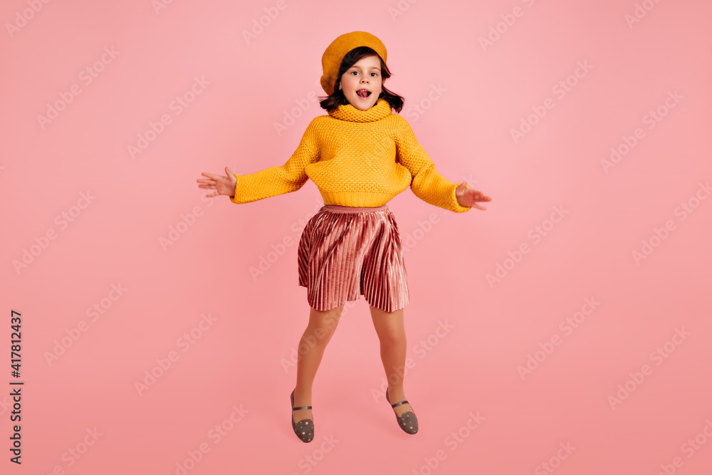 Pleased preteen girl in knitted sweater dancing on pink background. Studio shot of jumping child wears yellow beret. Pleased preteen girl in knitted sweater dancing on pink background. Studio shot of jumping child wears yellow beret.